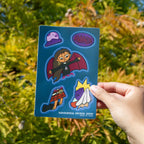 A hand holding a sticker sheet of a Filipino mythical creature called Manananggal with a moon and a cloud and garlic and salt against a leafy background