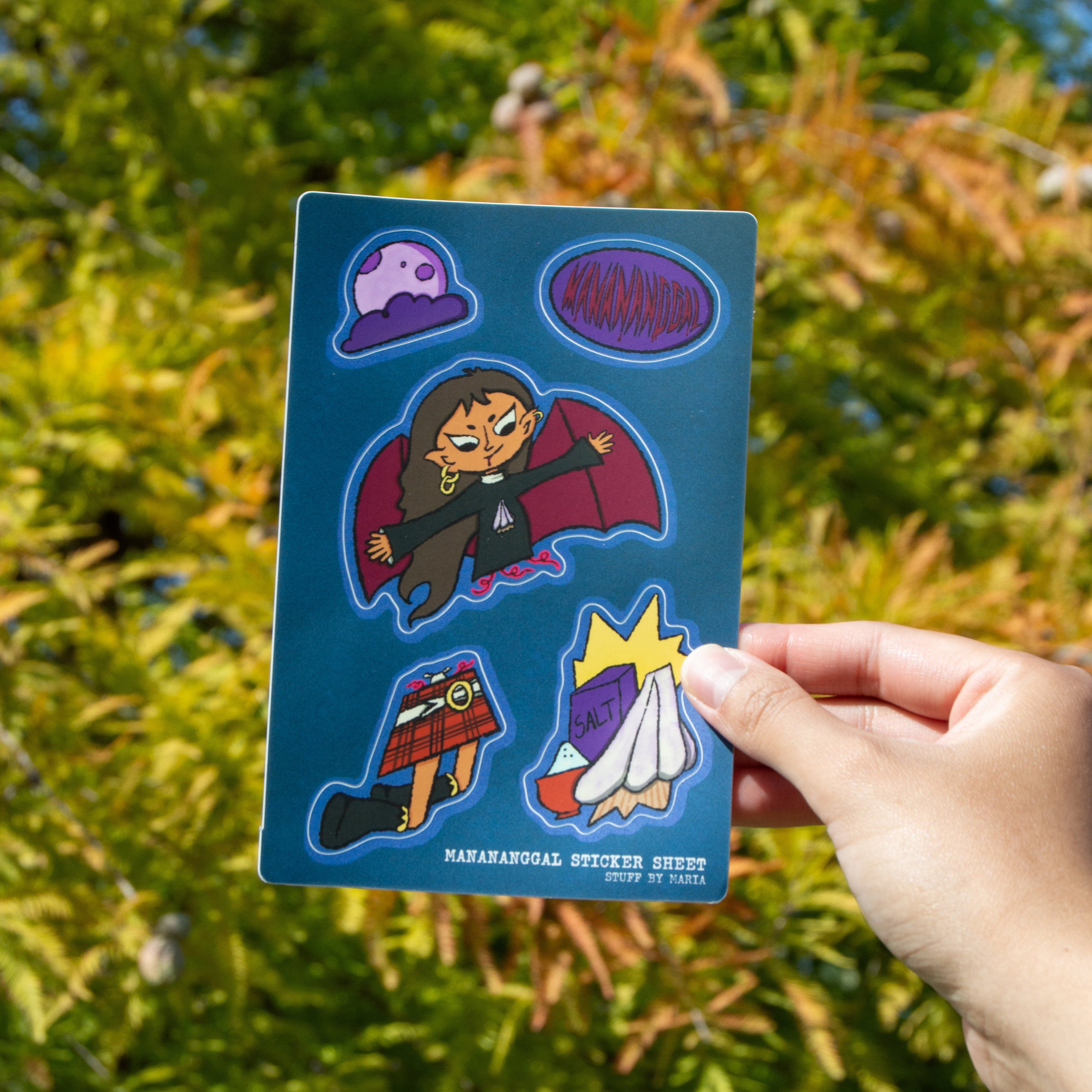 A hand holding a sticker sheet of a Filipino mythical creature called Manananggal with a moon and a cloud and garlic and salt against a leafy background